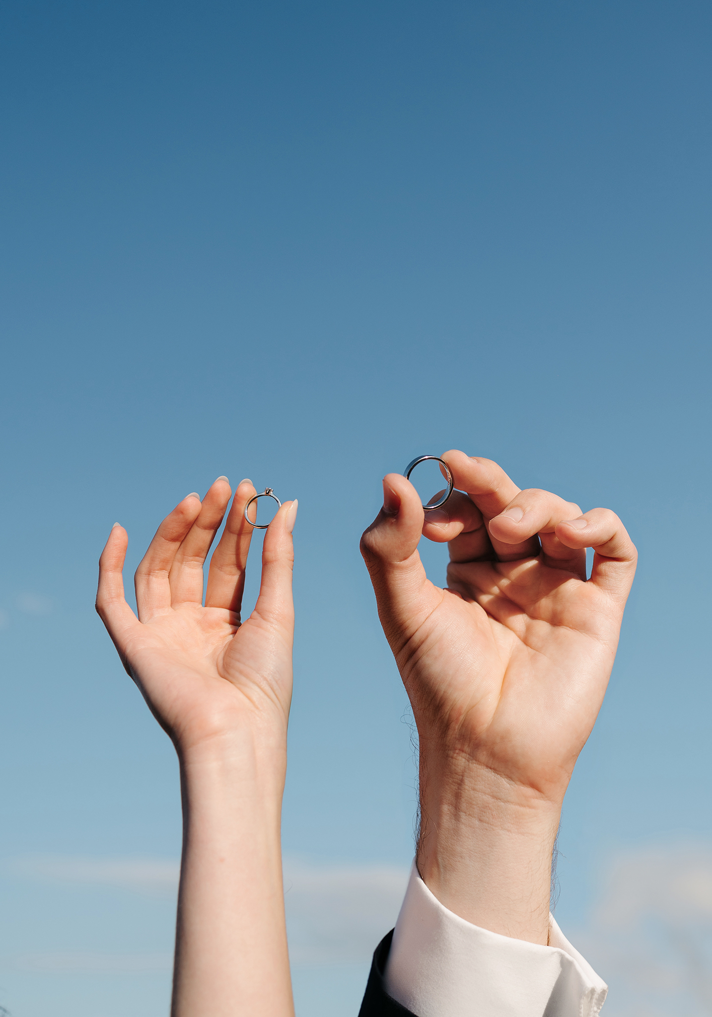 bride and groom holding their wedding bands up in the air with blue sky in the background
